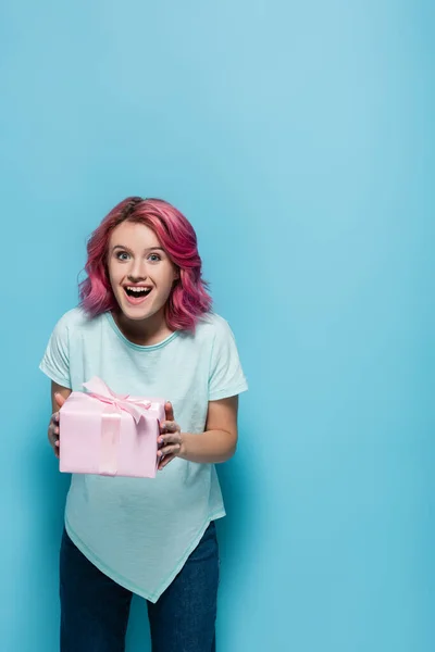 Aufgeregte junge Frau mit rosa Haaren hält Geschenkbox mit Schleife auf blauem Hintergrund — Stockfoto