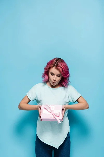 Überrascht junge Frau mit rosa Haaren hält Geschenkbox mit Schleife auf blauem Hintergrund — Stockfoto