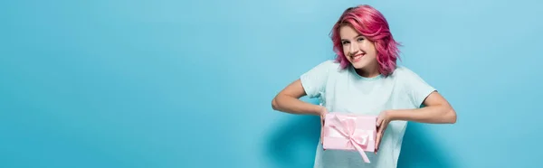 Jovem mulher com cabelo rosa segurando caixa de presente com arco e sorrindo no fundo azul, tiro panorâmico — Fotografia de Stock