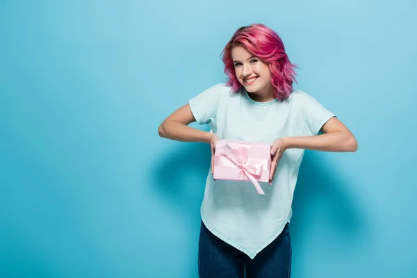 Jovem mulher com cabelo rosa segurando caixa de presente com arco e sorrindo no fundo azul — Fotografia de Stock
