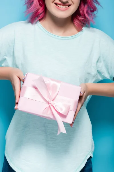 Vista recortada de mujer joven con pelo rosa sosteniendo caja de regalo con arco y sonriendo sobre fondo azul - foto de stock