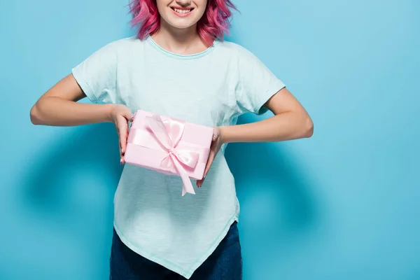 Обрезанный вид молодой женщины с розовыми волосами проведение подарочную коробку с луком и улыбаясь на синем фоне — стоковое фото