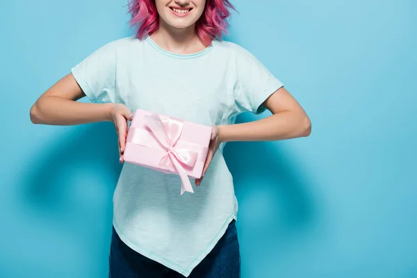 Cropped view of young woman with pink hair holding gift box with bow and smiling on blue background — Stock Photo