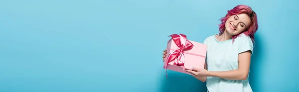 Young woman with pink hair holding gift box with bow and smiling with closed eyes on blue background, panoramic shot — Stock Photo