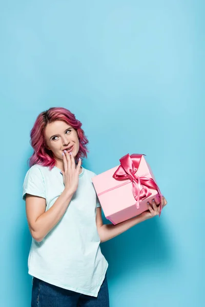 Schüchterne junge Frau mit rosa Haaren hält Geschenkbox mit Schleife auf blauem Hintergrund — Stockfoto
