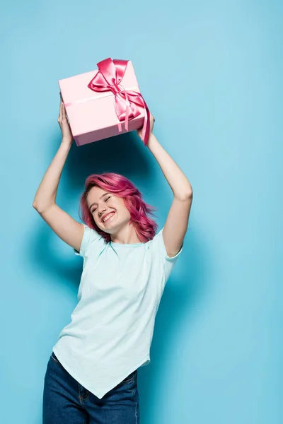 Young woman with pink hair holding gift box with bow above head and smiling on blue background — Stock Photo