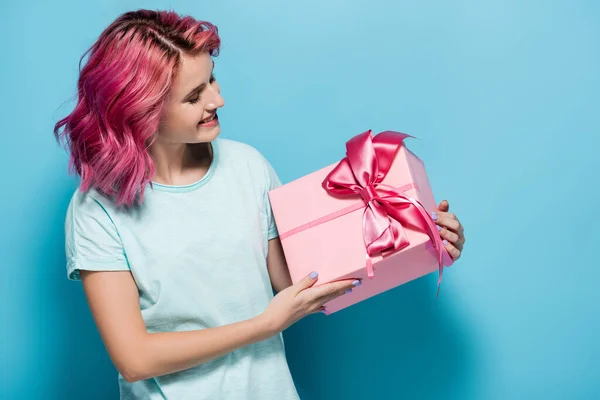 Молода жінка з рожевим волоссям дивиться на подарункову коробку з бантом і посміхається на синьому фоні — стокове фото