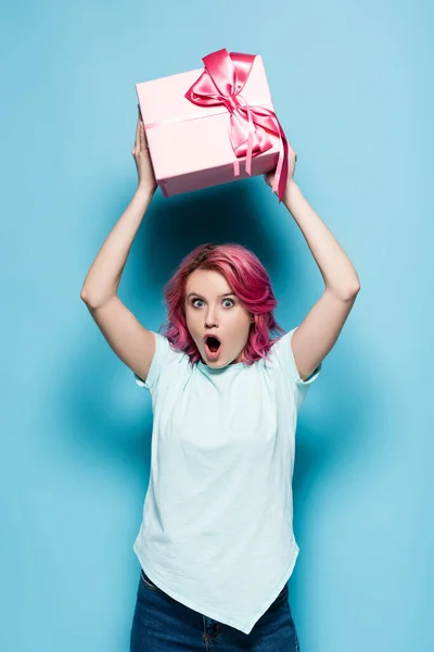 Schockierte junge Frau mit rosa Haaren und offenem Mund hält Geschenkbox mit Schleife auf blauem Hintergrund — Stockfoto