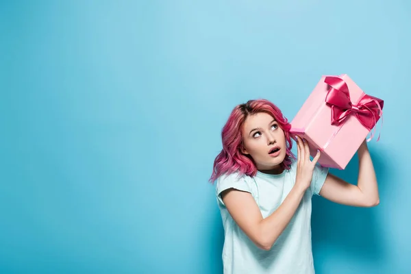 Young woman with pink hair and open mouth holding gift box with bow on blue background — Stock Photo