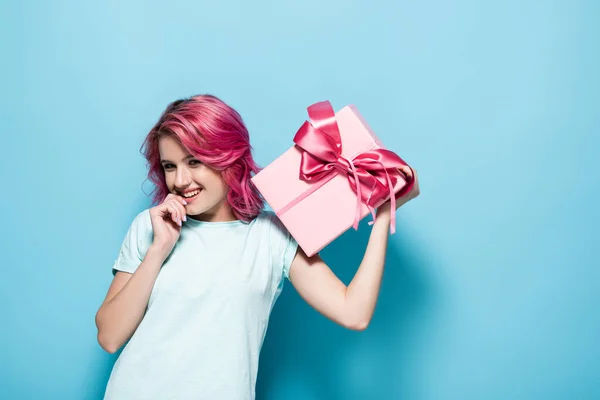 Young woman with pink hair holding gift box with bow and smiling on blue background — Stock Photo