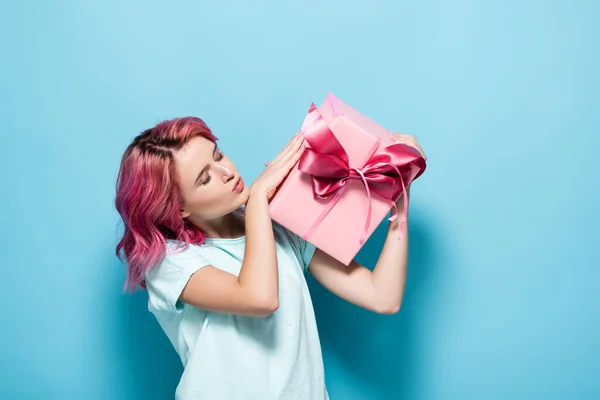 Young woman with pink hair kissing gift box with bow on blue background — Stock Photo