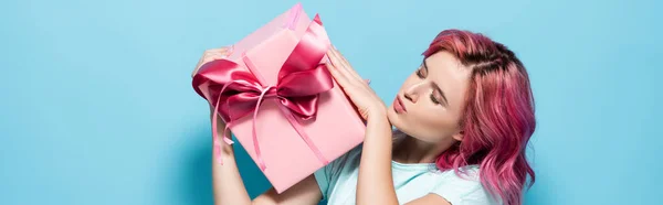 Young woman with pink hair kissing gift box with bow on blue background, panoramic shot — Stock Photo