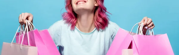 Vista recortada de mujer joven con el pelo rosa sosteniendo bolsas de compras y sonriendo aislado en azul - foto de stock