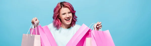 Excitée jeune femme aux cheveux roses tenant des sacs à provisions isolés sur bleu, plan panoramique — Photo de stock