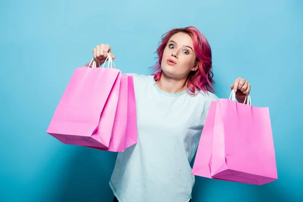 Schockierte junge Frau mit rosa Haaren, die Einkaufstüten auf blauem Hintergrund hält — Stockfoto