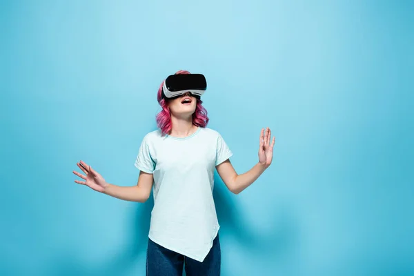 Schockierte junge Frau mit rosa Haaren im vr-Headset gestikuliert auf blauem Hintergrund — Stockfoto