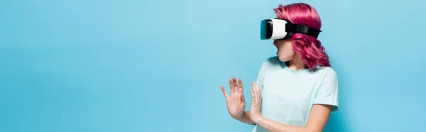 Scared young woman with pink hair in vr headset gesturing on blue background, panoramic shot — Stock Photo