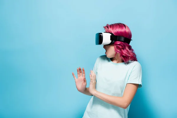 Spaventata giovane donna con i capelli rosa in auricolare vr gesticolare su sfondo blu — Foto stock