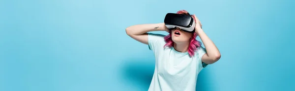 Impactado mujer joven con el pelo rosa en auriculares vr celebración de la cabeza sobre fondo azul, tiro panorámico - foto de stock