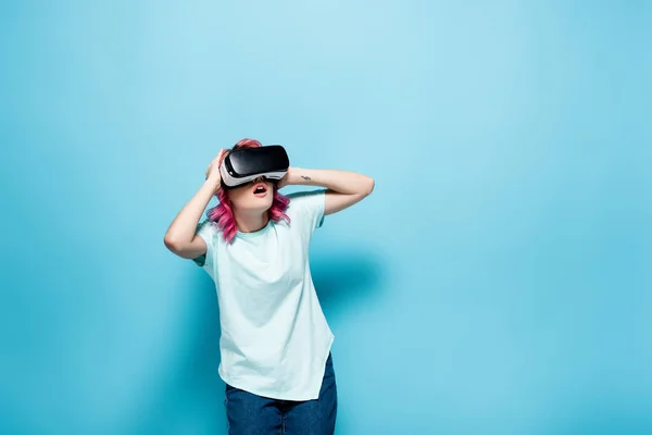 Mujer joven conmocionada con el pelo rosa en auriculares vr con la cabeza sobre fondo azul - foto de stock