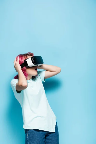 Mujer joven conmocionada con el pelo rosa en auriculares vr con la cabeza sobre fondo azul - foto de stock