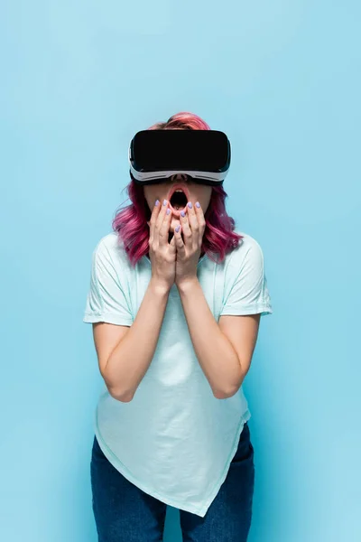 Choqué jeune femme avec des cheveux roses en vr casque toucher le visage sur fond bleu — Photo de stock