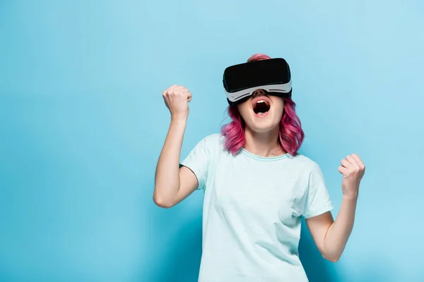 Возбужденная молодая женщина с розовыми волосами в VR гарнитуре показывая да жест на голубом фоне — стоковое фото