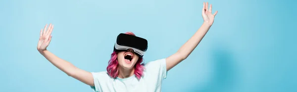 Excited young woman with pink hair in vr headset with hands in air on blue background, panoramic shot — Stock Photo