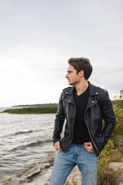 Young man in jeans and leather jacket looking away on sea coast — Stock Photo