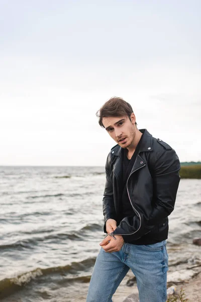Молодой человек в джинсах и кожаной куртке смотрит в камеру на пляже — стоковое фото