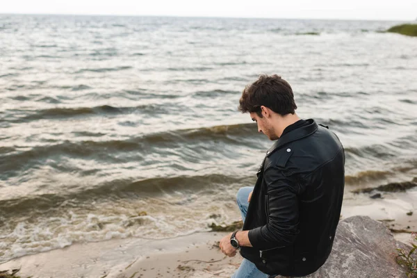 Man in leather jacket sitting on stone on sea coast — Stock Photo