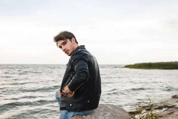 Young man in leather jacket looking at camera on stone near sea — Stock Photo
