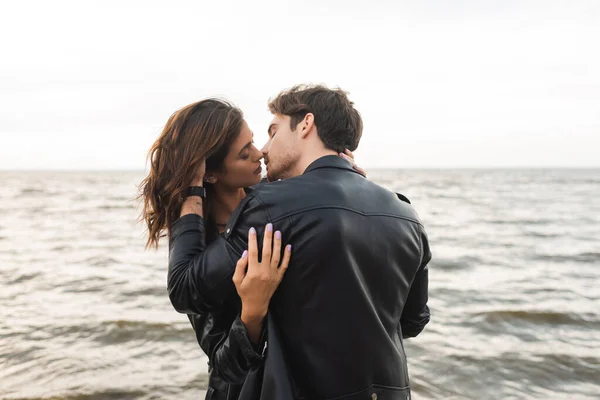Man in leather jacket kissing girlfriend with sea at background — Stock Photo