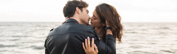 Vue panoramique de l'homme en veste en cuir embrasser et toucher les cheveux de la petite amie avec la mer à l'arrière-plan — Photo de stock