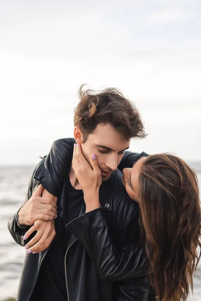 Young woman touching cheek and embracing boyfriend in leather jacket near sea — Stock Photo