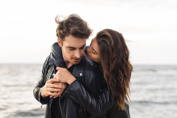 Young brunette woman hugging boyfriend in black leather jacket near sea — Stock Photo