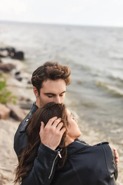 Young man touching hair of brunette girlfriend in leather jacket near sea — Stock Photo