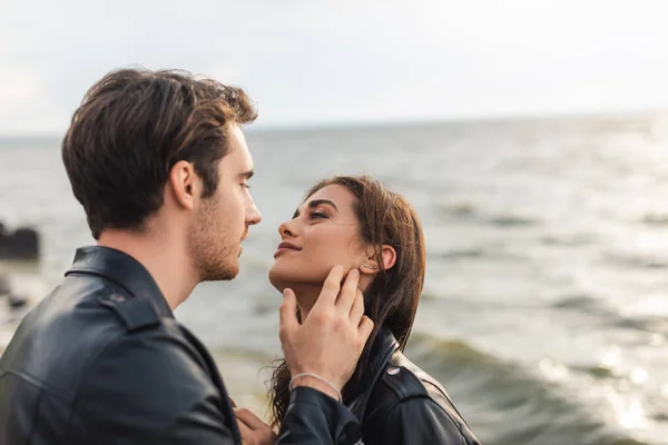 Young man touching cheek of brunette girlfriend in leather jacket on beach — Stock Photo