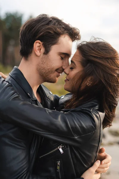 Young man kissing brunette girlfriend in leather jacket outdoor — Stock Photo