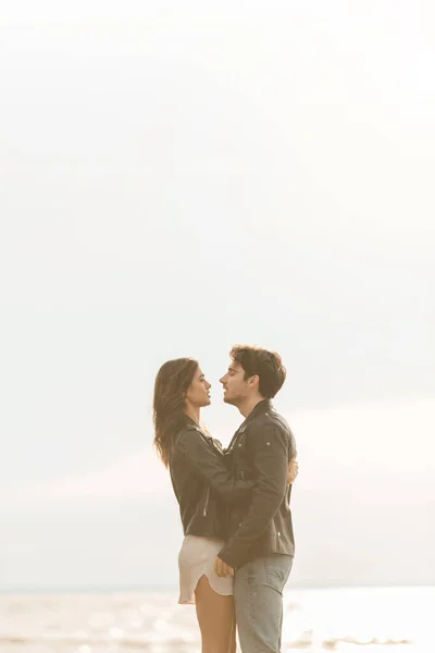 Side view of woman in leather jacket hugging boyfriend with sea and sky at background — Stock Photo