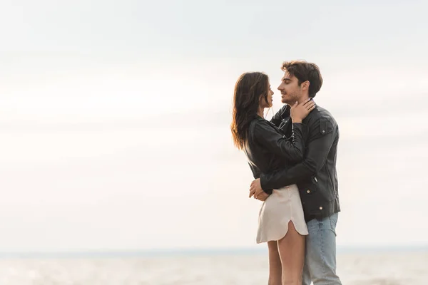 Young man hugging brunette girlfriend in leather jacket with sky and sea at background — Stock Photo