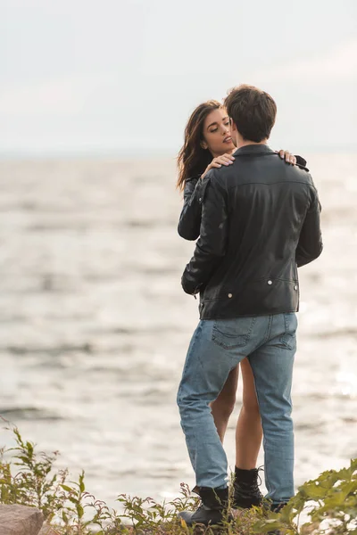 Mulher morena abraçando namorado em jaqueta de couro e jeans na praia perto do mar — Fotografia de Stock