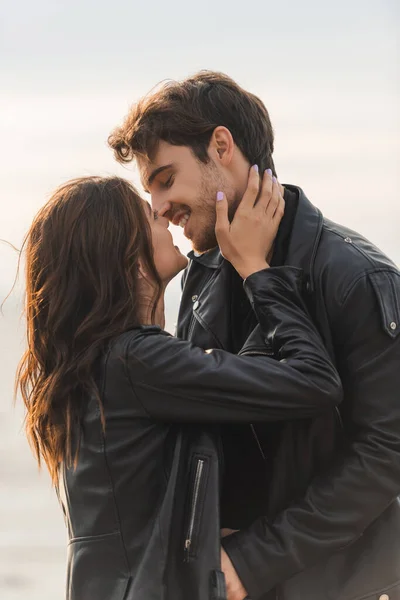 Mujer morena besando y tocando novio en chaqueta de cuero al aire libre - foto de stock