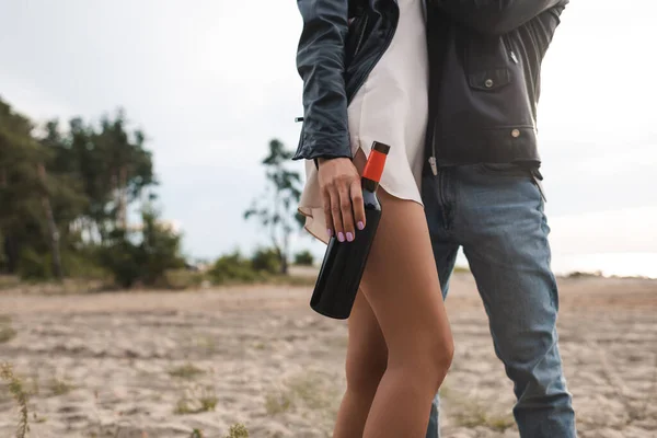 Vista ritagliata della donna in abito e giacca di pelle in possesso di bottiglia di vino vicino fidanzato sulla spiaggia — Foto stock