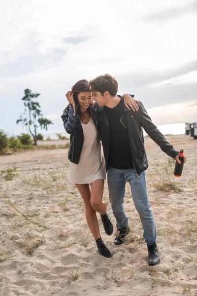 Uomo in giacca di pelle abbracciando fidanzata e tenendo bottiglia di vino sulla spiaggia — Foto stock