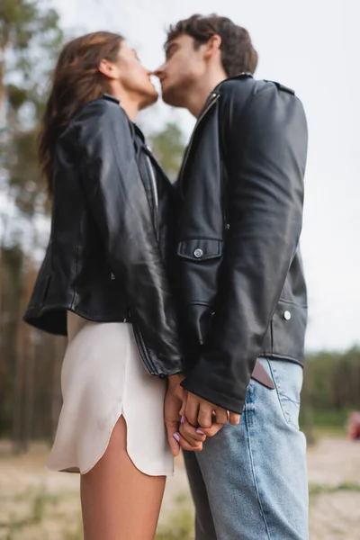 Low angle view of young couple in leather jackets holding hands and kissing in forest — Stock Photo