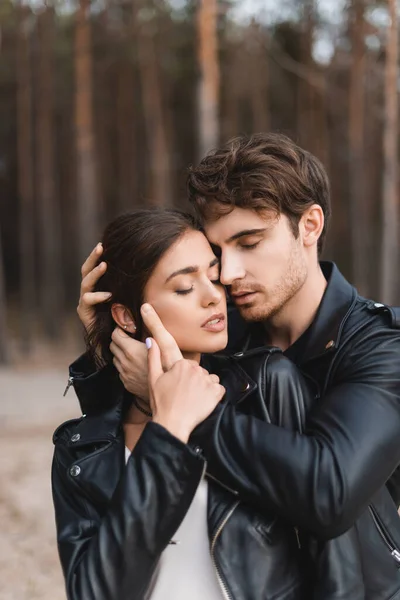 Young man with closed eyes embracing brunette girlfriend in leather jacket near forest — Stock Photo