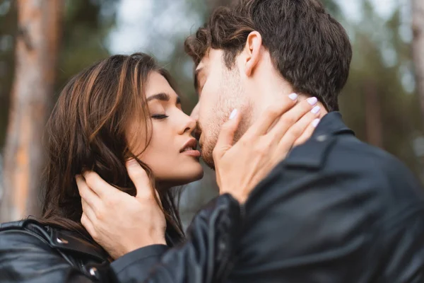 Foco seletivo de jovem casal em jaquetas de couro beijando na floresta — Fotografia de Stock