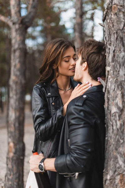 Selective focus of woman kissing boyfriend in leather jacket with bottle of wine in forest — Stock Photo