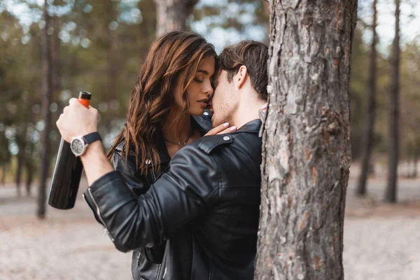 Selective focus of man in leather jacket kissing girlfriend and holding bottle of wine in forest — Stock Photo